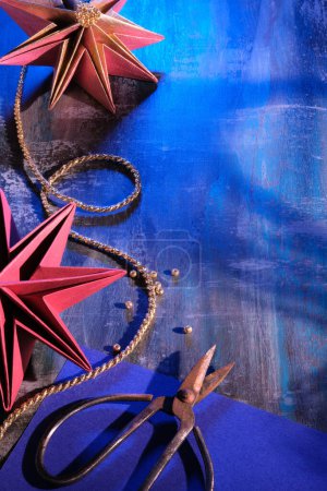 Foto de Estrellas de papel hechas a mano, decoraciones navideñas hechas a mano. Adornos de Navidad sobre fondo oscuro. Origami hobby artesanal. Tijeras, cordón dorado y purpurina sobre mesa de madera oscura, espacio-copia.. - Imagen libre de derechos