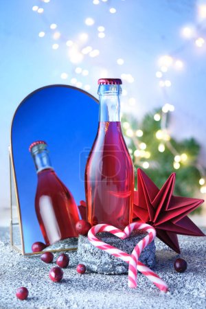 Photo for Small bottle of rose, pink vine on stone pedestal reflected in arc mirror. Christmas, wintertime celebration concept. Xmas wintertime decor, festive light garland, berry, paper stars on dark background. - Royalty Free Image