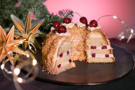 Foto de Pastel alemán Frankfurter Kranz o Frankfurt Crown Cake. Bisquit con crema de mantequilla, cereza. La mitad del pastel en el plato negro. Ramitas de abeto de Navidad, estrella de papel, guirnalda con luces. - Imagen libre de derechos