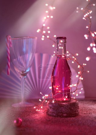 Photo for Small bottle of rose, pink vine or cranberry vodka on stone pedestal. Christmas celebration concept. Drinking glass and Xmas wintertime decor, festive light garland, paper fan on dark background. - Royalty Free Image