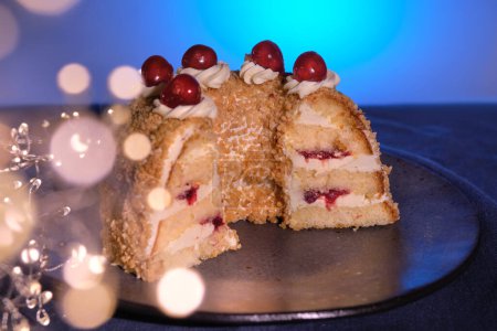 Foto de Pastel alemán Frankfurter Kranz o Frankfurt Crown Cake. Bisquit con crema de mantequilla y cereza. Corta la mitad del pastel. Guirnalda de Navidad de invierno con luces festivas. Mantel azul oscuro, fondo azul. - Imagen libre de derechos