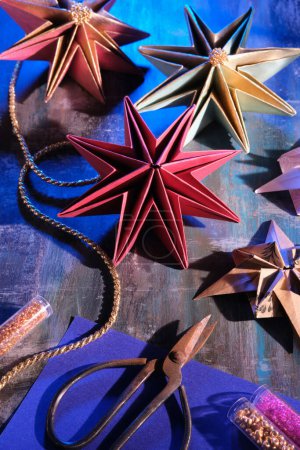 Foto de Estrellas de papel hechas a mano, decoraciones navideñas hechas a mano. Adornos de Navidad sobre fondo oscuro. Origami hobby artesanal. Tijeras, cordón dorado y purpurina sobre mesa de madera oscura. - Imagen libre de derechos