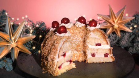 Foto de Pastel alemán Frankfurter Kranz o Frankfurt Crown Cake. Bisquit con crema de mantequilla, cereza. La mitad del pastel en el plato negro. Ramitas de abeto de Navidad, estrella de papel, guirnalda con luces. - Imagen libre de derechos