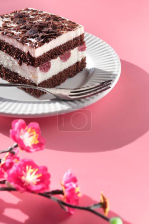 Téléchargez les photos : Gâteau au chocolat aux cerises acides. Pièce de gâteau sur une assiette avec fourchette. Dessert sucré sur fond rose avec des fleurs de prunes magenta vibrantes. Tranche de torte avec des fleurs de printemps sur les brindilles. - en image libre de droit