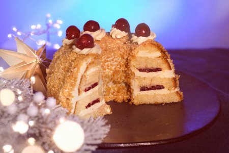 Foto de Pastel alemán Frankfurter Kranz o Frankfurt Crown Cake. Bisquit con crema de mantequilla y cereza. Corta la mitad del pastel en un plato negro. Ramitas de abeto de Navidad, estrella de papel, guirnalda con luces. - Imagen libre de derechos