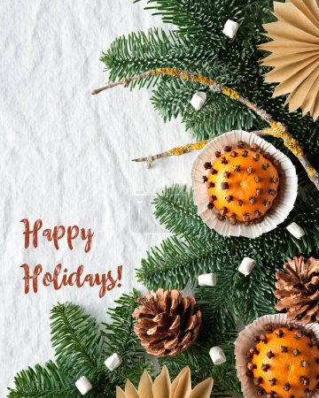 Photo for Fragrant pomander balls made with tangerines with cloves. Text, caption, greeting Happy Holidays on tablecloth. Paper stars from brown paper. Flat lay on off white textile and decorated fir twigs. - Royalty Free Image