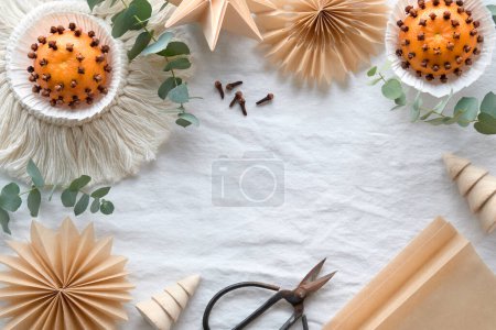 Photo for Fragrant pomander balls handmade from tangerines with cloves. Flat lay on off white textile tablecloth with aromatic wintertime green eucalyptus twigs. Old scissors and brown paper stars, snowflakes. - Royalty Free Image