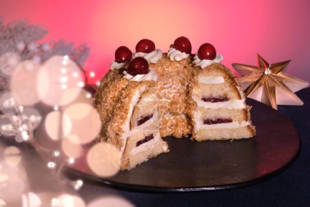 Photo for German cake Frankfurter Kranz or Frankfurt Crown Cake. Bisquit with butter cream, cherry. Half of the cake on black plate. Black and pink background.Xmas fir twigs, paper star, garland with lights. - Royalty Free Image