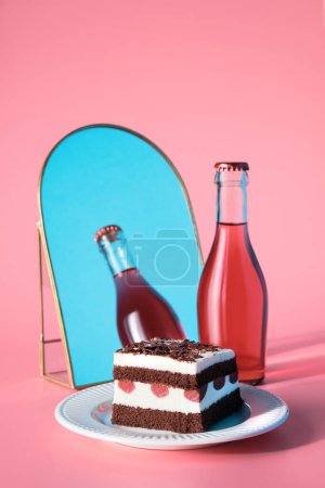 Téléchargez les photos : Gâteau au chocolat aux cerises acides. Pièce de gâteau sur une assiette avec fourchette. Sucré dessert sur fond rose. Vin rose en petite bouteille, ciel bleu reflété dans le miroir voûte. - en image libre de droit