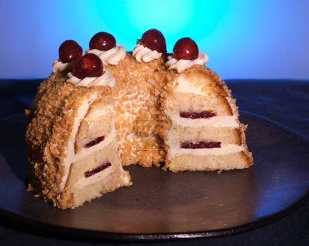 Foto de Pastel alemán Frankfurter Kranz o Frankfurt Crown Cake. Bisquit con crema de mantequilla y cereza. Corta la mitad del pastel en un plato negro. Mantel azul oscuro, fondo azul. - Imagen libre de derechos