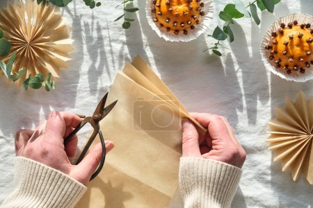 Photo for Fragrant pomander balls handmade from tangerines and cloves. Flat lay on off white textile tablecloth with aromatic wintertime green eucalyptus twigs. Hands making paper stars from brown baking paper. - Royalty Free Image