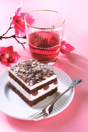 Photo for Chocolate cake with sour cherries. Piece of cake on a plate with fork. Sweet dessert on pink background with Spring magnolia blossoms, pink flower decorations. Glass of pink lemonade or rose wine. - Royalty Free Image