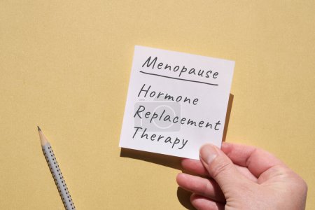 Photo for Hand hold paper page with text Menopause, hormone replacement therapy. Top view, flat lay on yellow monochromatic paper background with pencil. Writing on blank square post it note. - Royalty Free Image