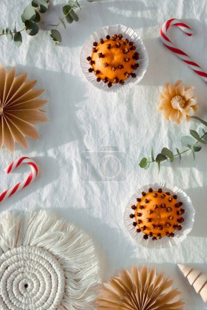 Photo for Fragrant pomander balls handmade from tangerines with cloves. Handmade paper stars from brown baking paper. Flat lay on off white textile tablecloth, eucalyptus. Wood Christmas tree toys, candy canes. - Royalty Free Image