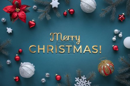 Photo for Text Merry Christmas. Festive Xmas decorations on dark turquoise emerald green textile tablecloth. Shiny golden greeting caption. Fir twigs, poinsettia flower, white and red toys, snowflakes and mirror disco balls. - Royalty Free Image