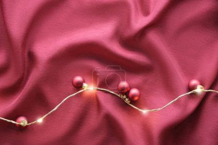 Photo for Viva Magenta color of the year 2023. Dark red Christmas background. Monochromatic flat lay with Xmas baubles and garland of electric festive lights on red silk textile background. - Royalty Free Image