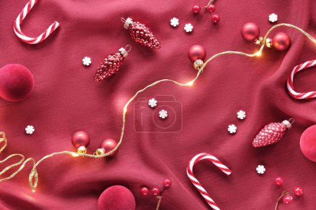 Photo for Viva Magenta color of the year 2023. Dark red Christmas background. Monochromatic flat lay with Xmas baubles, candy canes, and garland of electric festive lights on red silk textile background. - Royalty Free Image