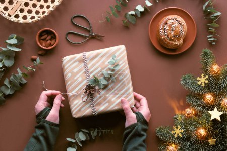 Téléchargez les photos : Fond de Noël brun avec des mains enveloppant boîte cadeau de Noël. Pose plate avec des bonbons de massepain, boîtes-cadeaux enveloppées, eucalyptus, étoiles, pain à la cannelle. Vue de dessus, sapin avec flocons de neige et guirlande électrique. - en image libre de droit