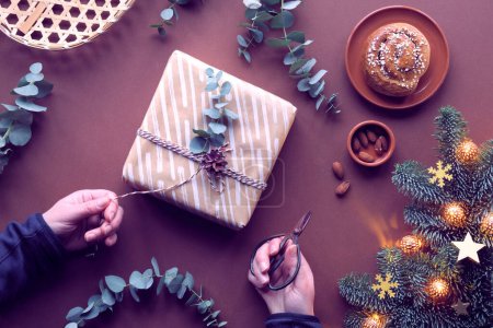 Photo for Chestnut brown Christmas background. Hands wrapping Xmas gift box. Flat lay with marzipan sweets, wrapped gift boxes, eucalyptus, stars, cinnamon bun. Fir twig with snowflakes and electric garland. - Royalty Free Image