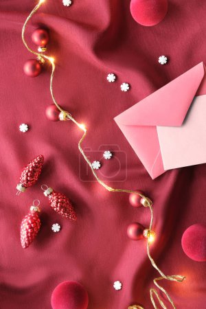 Photo for Viva Magenta color of the year 2023. Dark red Christmas background with paper card, envelope. Monochrome flat lay. Xmas baubles, candy canes, and garland with festive lights on red silk textile. - Royalty Free Image