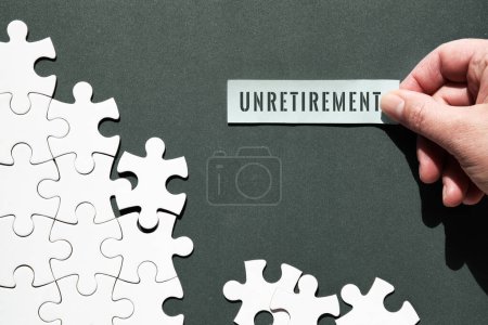 Photo for Unretirement concept image. Puzzle pieces half assembled and separate. Hand holding piece of paper, sticky note with word Unretirement on it. Top view, flat lay on dark green paper background. - Royalty Free Image