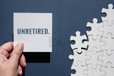 Photo for Unretired word written on square paper note in hand. Top view, flat lay on dark blue monochromatic paper background with assembled white puzzle pieces. - Royalty Free Image