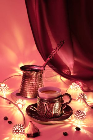 Téléchargez les photos : Cafetière turque et tasse traditionnelle avec des bonbons orientaux. Guirlande métallique en cuivre ornée et lampes à thé. Fond de fête monochromatique rouge vif et doré éclairé par des chandelles. - en image libre de droit