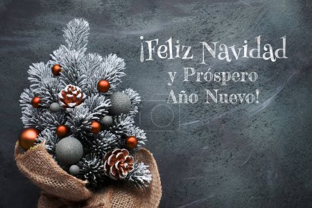 Photo for Small Christmas tree in sackcloth decorated with red baubles and berries on dark textured background. Feliz Navidad means Merry Christmas in Spanish language. - Royalty Free Image