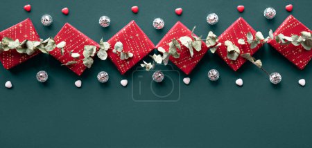 Téléchargez les photos : Wintertime green red decorative border with gift boxes, disco balls and eucalyptus. Flat lay on dark green, cyan linen textile. Panorama with wrapped presents and dry winter plants. Panoramic banner with border. - en image libre de droit