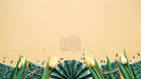 Téléchargez les photos : Decrative Spring border with yellow tulip flowers and gift boxes. Golden yellow paper background with presents in green wrapping paper and confetti. Flat lay, top view with copy-space, place for text. - en image libre de droit