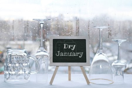 Téléchargez les photos : Dry January, month without alcohol. Chalk text, caption Dry January on blackboard, chalk board. Empty vine and beer drinking glasses. Window with raindrops, grey winter city skyline on background. - en image libre de droit