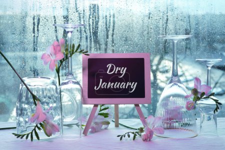 Foto de Dry January, month without alcohol. Text on blackboard, chalk board. Empty vine and beer drinking glasses, freesia flowers. Window with raindrops on background. - Imagen libre de derechos