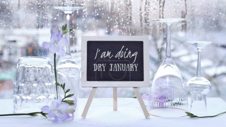 Foto de Dry January, month without alcohol. Text I am doing dry January on blackboard, chalk board. Empty vine and beer drinking glasses, freesia flowers. Window with raindrops grey winter day on background. - Imagen libre de derechos