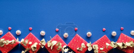 Téléchargez les photos : Wintertime blue red decorative border with gift boxes, disco balls and eucalyptus. Flat lay on blue linen textile. Panorama with wrapped presents and dry winter plants. Panoramic banner with border. - en image libre de droit