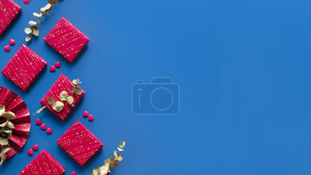 Photo for Wintertime magenta decor, gift boxes. Flat lay on blue linen textile, copy-space, text place. Panorama with wrapped presents, paper fan, eucalyptus. Red blue arrangement, panoramic banner composition. - Royalty Free Image