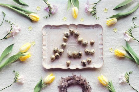 Foto de Natural Easter decor, overhead, flat lay with quail eggs, feather wreath, yellow tulip flowers on off white textile background. Flat lay, top view natural low impact springtime background.. - Imagen libre de derechos