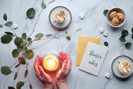 Foto de Card with greeting winter vibes. Hands around warm candle. Mint green postal envelope, blank white card, coffee cups and cookies on table with eucalyptus. - Imagen libre de derechos
