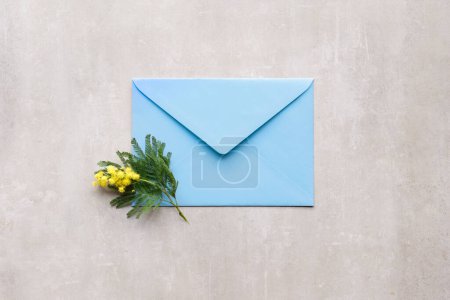 Photo for Mimosa branch with flowers and blue envelope on beige natural stone background. Natural springtime decor. Flat lay, top view. - Royalty Free Image