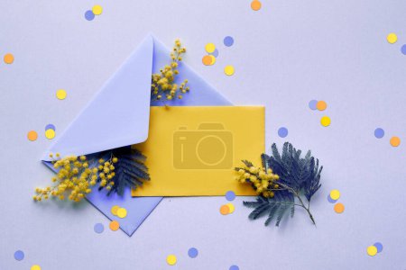 Photo for Blank yellow paper card, fresh mimosa flowers in pale violet envelope on lilac background with paper confetti. Natural springtime decor in two colors. Flat lay, top view, central composition. - Royalty Free Image