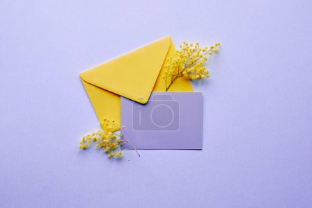 Photo for Blank pale violet paper card, yellow mimosa flowers in envelope on lilac background. Natural springtime decor in two colors. Flat lay, top view, central composition, simple, minimal arrangement. - Royalty Free Image