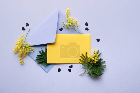 Foto de Blank yellow paper card, fresh mimosa flowers in pale violet envelope on lilac background with paper confetti. Natural springtime decor in two colors. Flat lay, top view, central composition. - Imagen libre de derechos
