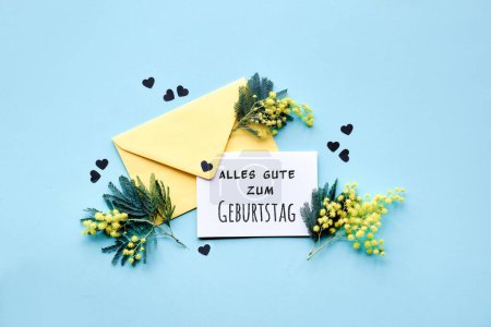 Téléchargez les photos : Texte Alles Gute zum Geburtstag signifie Joyeux anniversaire en langue allemande. Texte Moments spéciaux sur carte de papier avec des fleurs de mimosa dans une enveloppe jaune sur fond bleu menthe, confettis. Couché plat, vue du dessus. - en image libre de droit