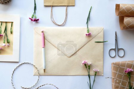 Téléchargez les photos : Enveloppe et concept de carte de vœux faite par soi-même avec des fleurs d'oeillet fraîches, style monticule. Cadeau enveloppé, carte postale en papier, enveloppe en papier, cordon, ciseaux et œillets violets. - en image libre de droit