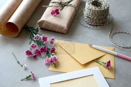 Photo for Envelope and self made greeting card concept with fresh carnation flowers. Wrapped gift, paper postcard, paper envelope, cord, scissors and purple carnations. - Royalty Free Image