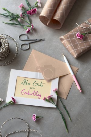 Photo for Envelope and self made greeting card concept with fresh carnation flowers. Wrapped gift, paper postcard, paper envelope. Text Alles Gute zum Geburtstag means Happy Birthday in German language.. - Royalty Free Image