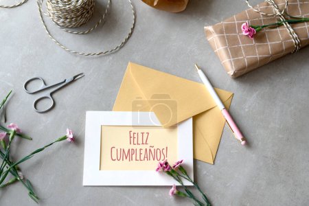Photo for Envelope and greeting card concept with carnation flowers. Wrapped gift, paper postcard, paper envelope, cord and scissors. Text Feliz Cumpleanos means Happy Birthday in Spanish language. - Royalty Free Image
