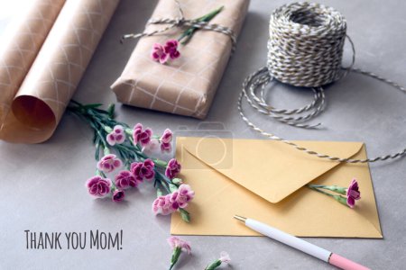 Téléchargez les photos : Enveloppe et carte de vœux de la fête des mères avec des fleurs fraîches d'oeillet. Cadeau enveloppé, carte postale en papier, enveloppe en papier, cordon, ciseaux et œillets violets. Légende Merci maman. - en image libre de droit