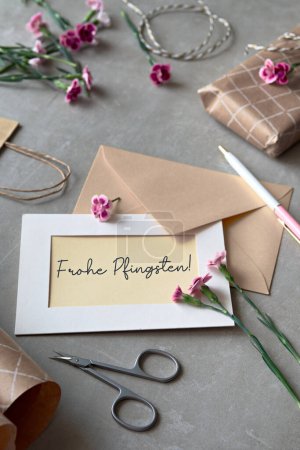 Photo for Text Frohe Phingsten means Happy Pentecost in German language. Envelope and greeting card with fresh carnation flowers. Wrapped gift, paper postcard, paper envelope, cord, and scissors on table. - Royalty Free Image
