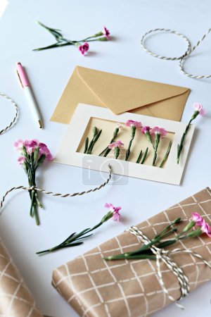 Téléchargez les photos : Enveloppe et concept de carte de vœux faite par soi-même avec des fleurs d'oeillet fraîches, style monticule. Cadeau enveloppé, carte postale en papier, enveloppe en papier, cordon, ciseaux et œillets violets. - en image libre de droit