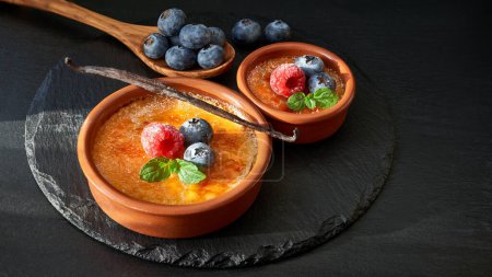 Photo for Creme brulee with raspberry, blueberry and mint leaves in terracota clay baking dishes on dark stone - Royalty Free Image